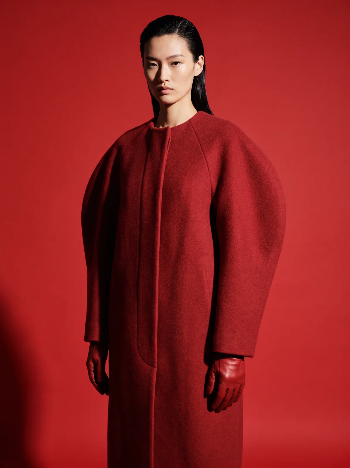 Manteau oversize sculptural en laine rouge garance, épaules arrondies, porté avec des gants en cuir rouge
