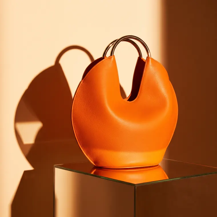 Sac à main sculptural en cuir orange mandarine, anse courbe en métal, posé sur un socle miroir